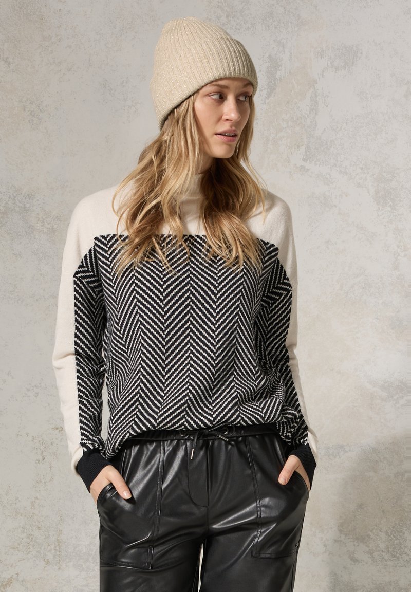 Maglione a righe herringbone in bianco e nero con maniche crema, abbinato a pantaloni in pelle nera larghi e un berretto beige a coste.