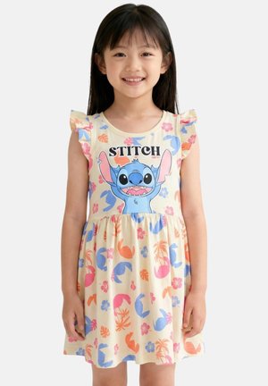 Fille souriante aux cheveux noirs et raides portant une robe crème avec des motifs tropicaux colorés et un grand personnage Stitch avec un texte sur la poitrine.