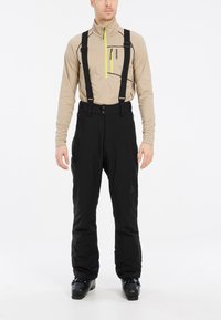 Pantalones de esquí negros con tirantes ajustables, bolsillos con cremallera y dobladillos reforzados. Llevados sobre una camiseta de manga larga beige.