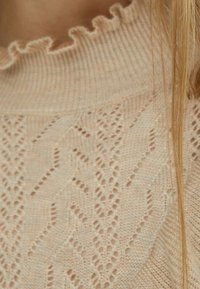 Lichtroze gebreide trui met een ruches aan de halslijn en een ingewikkeld, gestructureerd patroon met chevron- en ajourdetails.