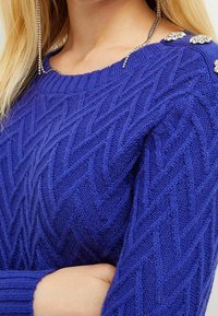Suéter de punto azul royal con un patrón de chevrón texturizado, con acentos de joyas en el hombro para un detalle adicional.