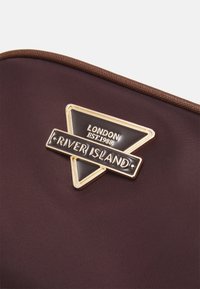 Hnědá nylonová taška s zlatou trojúhelníkovou nášivkou s černými akcenty, na které je embosovaný text "RIVER ISLAND" a "LONDÝN EST.1988."