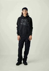 Young person wearing a black beanie, black graphic hoodie, black cargo pants, and black sneakers, standing against a plain light background.