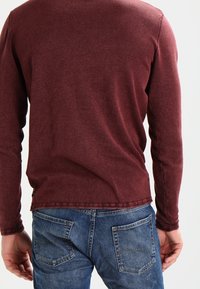 Chemise manches longues bordeaux avec un col rond, tissu texturé et ourlet droit, associée à un jean denim bleu avec des poches arrière.
