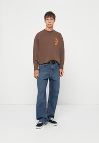 Ung mand står op, iført en brun langærmet skjorte med orange grafik, løse blå jeans med sort bælte og sorte sneakers med hvide såler.