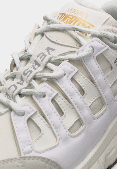 Gros plan sur une sneaker blanche Versace avec un design en couches de dentelle, un logo gris et doré, et une semelle épaisse blanche.