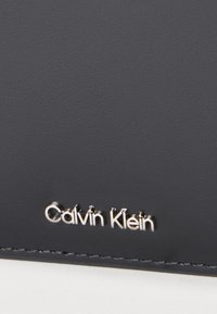 Μαύρο δερμάτινο με υφή και ανάγλυφο ασημένιο λογότυπο "Calvin Klein" στο κάτω μέρος. Η επιφάνεια είναι λεία, παρουσιάζοντας έναν μίνιμαλ σχεδιασμό.
