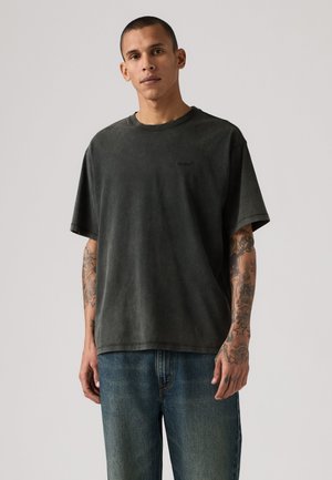 Homme aux cheveux courts portant un t-shirt noir délavé oversize et un jean bleu, montrant des tatouages sur les deux avant-bras sur un fond uni.