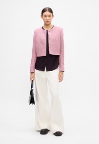 Jachetă roz scurtă cu trei butoane aurii, asortată cu un top închis la culoare și pantaloni albi, evazați. Modelul ține o geantă neagră, stând în fața unui fundal alb.