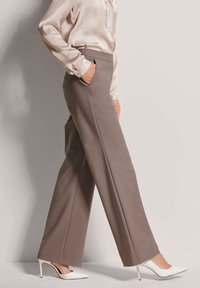 Op maat gemaakte beige broeken met een gladde textuur, hoge taille en zijzakken, gecombineerd met witte puntige hakken en een lichte blouse.