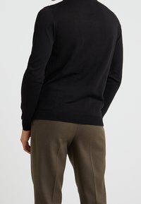 Schwarzer gestrickter Pullover mit Rundhalsausschnitt, enganliegender Silhouette und geripptem Saum, kombiniert mit olivgrünen, taillierten Hosen.