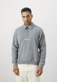 Γκρι φλις πουλό�βερ με κλείσιμο φερμουάρ στον γιακά, με ραμμένο σχέδιο και λευκές λογότυπες "BARCA" και Nike στο στήθος. Μαλακή υφή με ριμπ μανσέτες.