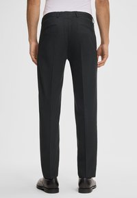 Zwarte broek met een gladde textuur, voorzien van een getailleerde pasvorm, een voornaad en twee achterzakken. Draagt met zwarte schoenen.