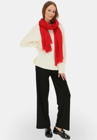 Kremowy oversize'owy sweter, czerwony szalik, czarne spodnie szerokie nogawki, czarne buty z błyszczącymi akcentami. Szalik ma frędzlowe brzegi.