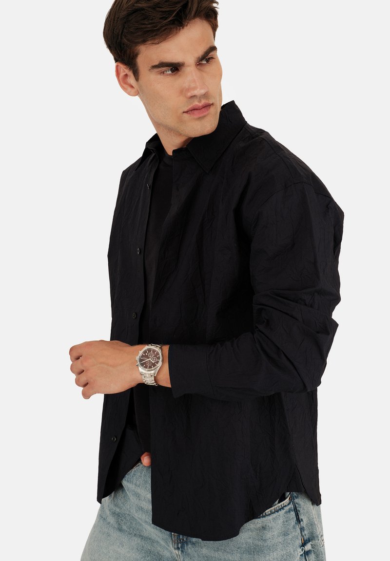 Chemise en tissu noir froissé avec un col boutonné, portée sur un top noir. Associée à une montre en argent, présentée sur un fond uni.