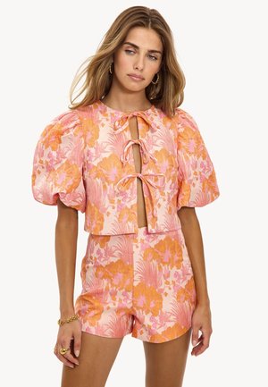 Vrouw in een roze en oranje bloemen tweedelige outfit met pofmouwen en een kortebroekset met gestrikte voorkant, geaccessorieerd met gouden sieraden.