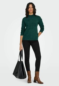 Tumši zaļa adīta džemperis ar augstu apkakli, kopā ar melnām plānām džinsa biksēm un brūnām papēžu zābakiem. Vienā rokā tiek turēta liela melna soma.