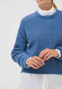 Modrý pletený svetr s texturovaným vzorem, kulatým výstřihem a dlouhými rukávy, navrstvený přes bílou košili s viditelnými manžetami.