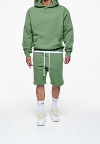 Sudadera verde con un bolsillo frontal, combinada con pantalones cortos verdes a juego con cordones. Zapatillas blancas y calcetines con acentos negros.