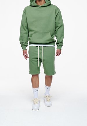 Felpa verde con tasca frontale, abbinata a pantaloni corti verdi coordinati con cordini. Sneaker bianche e calze con accenti neri.