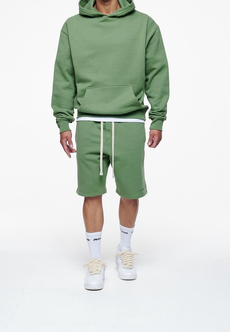 Sudadera verde con un bolsillo frontal, combinada con pantalones cortos verdes a juego con cordones. Zapatillas blancas y calcetines con acentos negros.