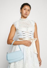 Ανοιχτό μπλε φόρεμα με λευκές δαντέλες, ζώνη με κορδέλα και λουλουδάτα μοτίβα. Αξεσουάρ περιλαμβάνουν μια μικρή ανοιχτό μπλε δερμάτινη τσάντα.