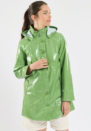 Femme portant un imperméable vert brillant avec capuche, debout devant un fond blanc uni, regardant sur le côté avec la main près de la poitrine.
