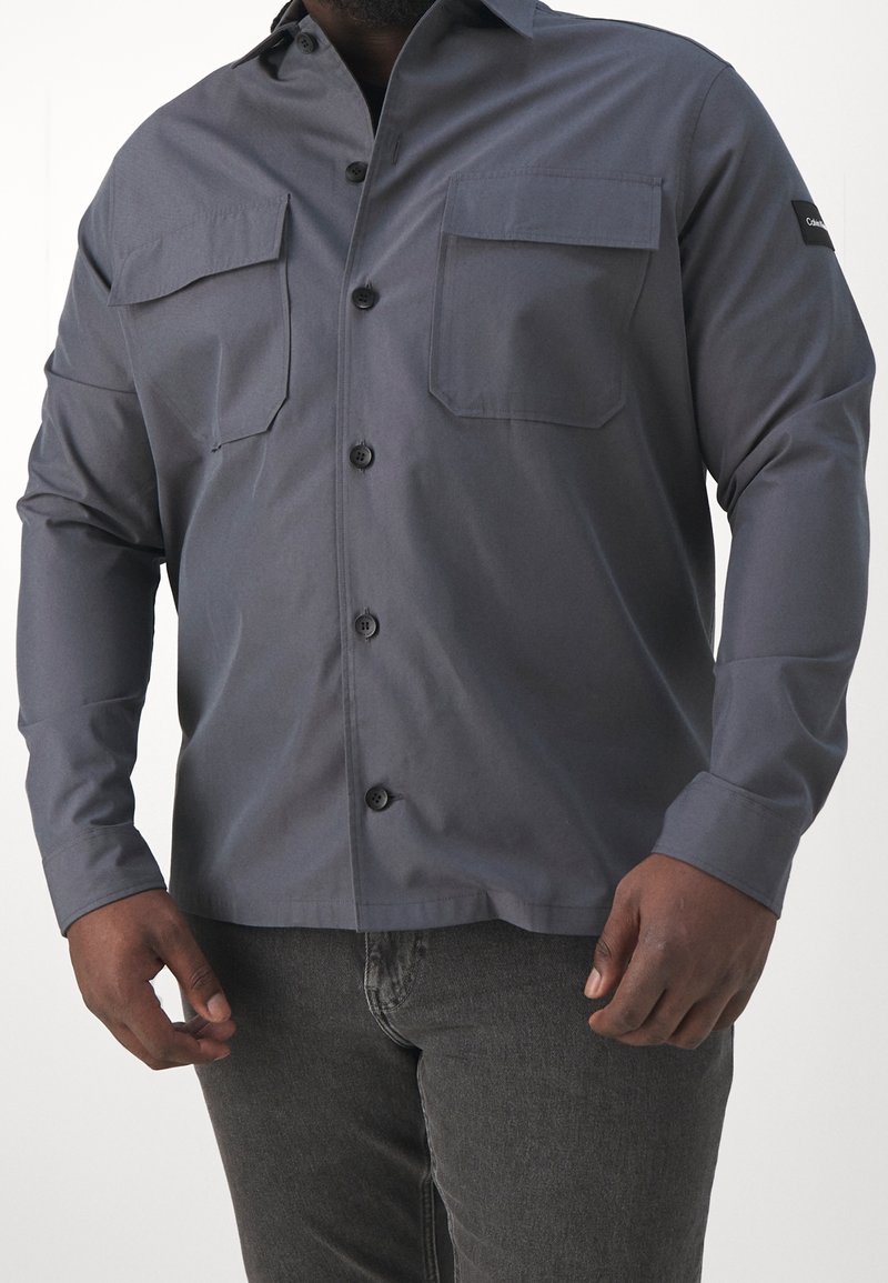 Homme portant une chemise boutonnée à manches longues gris foncé avec deux poches poitrine et un pantalon gris foncé, debout devant un fond uni.