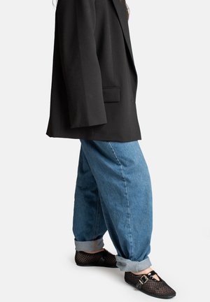 Persona vistiendo jeans azules holgados con los dobladillos enrollados, zapatos Mary Jane negros de malla y un blazer negro de gran tamaño, de pie contra un fondo liso.