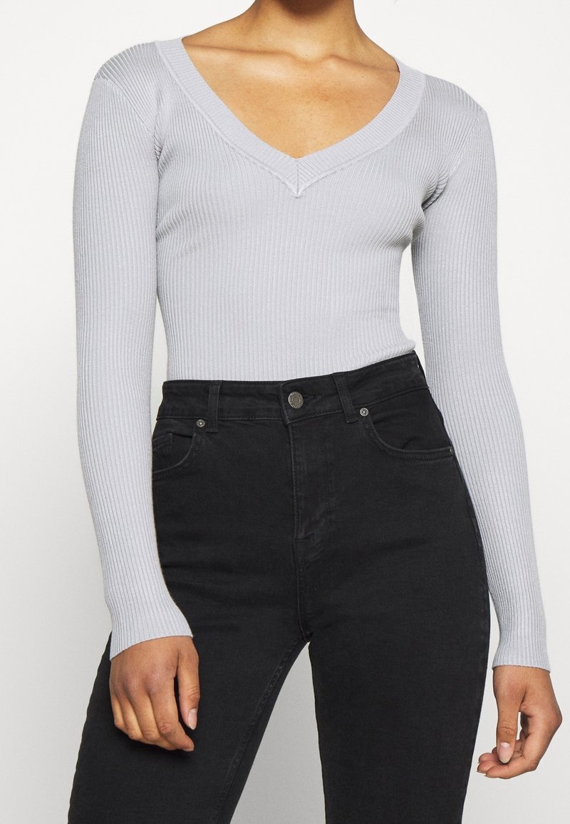 Femme portant un haut à manches longues gris clair côtelé avec col en V, rentré dans un jean noir taille haute, sur un fond uni.