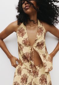 Světle žlutý halter top s květinovým potiskem v hnědých tónech, s hlubokým V výstřihem a zavazováním vpředu. Texturovaná látka, mírně volnější střih.