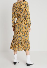 Vestido amarillo de flores de manga larga con acentos azules, dobladillo con volantes en capas y cintura ajustada, combinado con botas blancas hasta el tobillo.