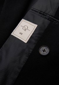 Chaqueta de tela negra con una textura suave, que presenta una etiqueta blanca con "Quintessence" y un botón negro con texto grabado.