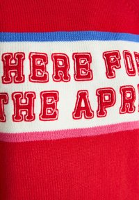 Red sweater featuring bold, red embroidered text that reads "HERE FOR THE APR" on a white background, accented with blue and pink stripes.