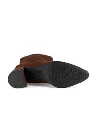 Bota de ante marrón para tobillo, tumbada de lado, mostrando la suela negra con un patrón ondulado y tacón de bloque sobre un fondo blanco.