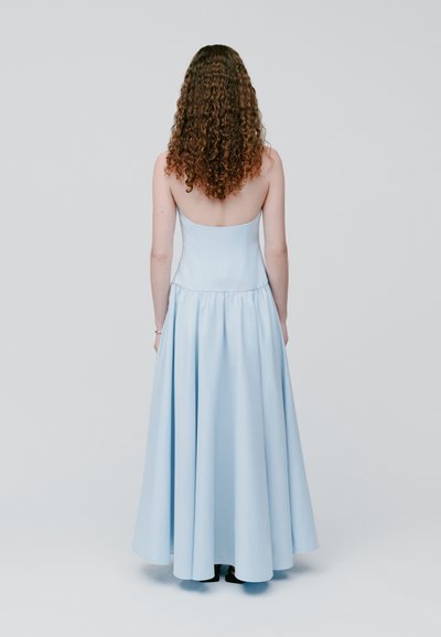 Γυναίκα με μακριά σγουρά μαλλιά, φορώντας ένα αμάνικο, μακρύ μέχρι το πάτωμα ανοιχτό μπλε φόρεμα, όρθια με την πλάτη στραμμένη προς έναν απλό φόντο.