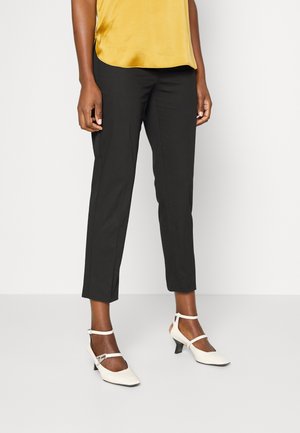 Persona con pantalones negros cortos, top sin mangas amarillo mostaza y zapatos blancos de tacón bajo con pulsera en el tobillo, de pie sobre un suelo blanco liso.
