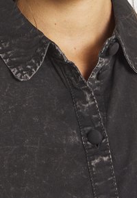 Czarna koszula na guziki z teksturowanym wzorem i kontrastującym błyszczącym kołnierzem. Posiada zaokrąglony kołnierz oraz zapięcie na guziki z przodu.