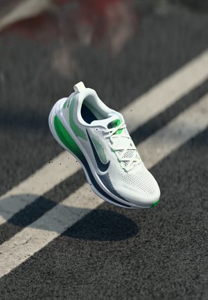Chaussure de course Nike blanche et verte en l'air au-dessus d'une route asphaltée avec des marquages de voie blancs, montrant les détails sur le côté et le dessus.