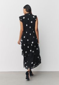 Abito lungo nero a pois con strati a balze, dettagli arricciati sulle spalle e vita aderente. Indossato con scarpe nere con tacco alto.