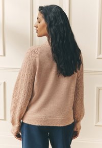 Jasnoróżowy sweter dziany z teksturowanym wzorem koronkowym na rękawach, zaokrąglonym dołem i miękkim materiałem. Noszony z ciemnoniebieskimi spodniami.