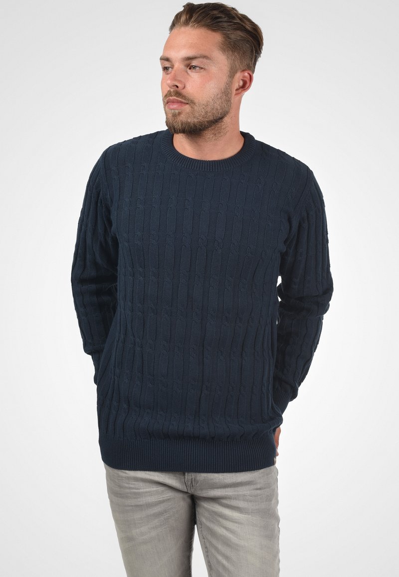 Pull en tricot bleu marine avec un motif de câbles texturé, col rond, et poignets et ourlet côtelés, porté avec un pantalon gris clair.