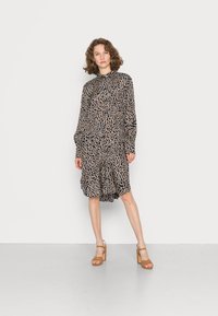 Vestido de mangas longas, com botões, na cor preta com um padrão abstrato em tom bege claro; tecido fluido, até ao joelho, combinado com sandálias de salto bloco em tom bege.