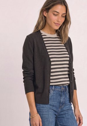 Femme portant un cardigan noir boutonné par-dessus une chemise beige et noire rayée avec un jean bleu, debout devant un fond clair uni.