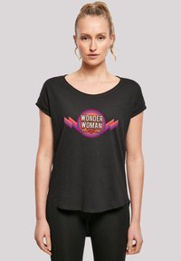 Melna īsām piedurknēm T-kreklu, kuram ir krāsains "Wonder Woman" grafiskais attēls ar apaļiem akcentiem, attēlota uz modeļa uz neitrāla fona.