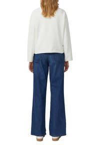 Biała, teksturowana bluzka z długim rękawem w połączeniu z ciemnoniebieskimi, szerokimi spodniami typu dżinsy z klasycznym pięciokieszeniowym krojem i widocznymi szwami.