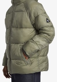 Puffjacka i olivgrön, med dragkedja, sidficka, vadderad struktur och svart logotypdetalj på ärmen.