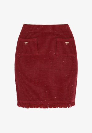 Raudona megzta sijonas su briaunotu paviršiumi, dviem priekinėmis kišenėmis, auksiniais kaspino akcentais ir plunksnuotu apvadu. Yra subtilių blizgučių elementų visame sijone.