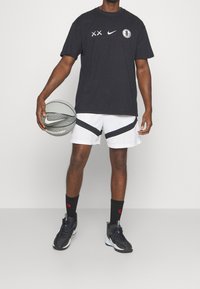 Camiseta negra de Nike con logo, pantalones cortos blancos con acentos diagonales negros, zapatos deportivos negros, sosteniendo un balón de baloncesto plateado de Nike.