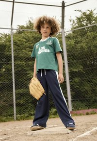 Grøn kortærmet skjorte med teksten "Los Angeles", parret med navy blå træningsbukser med hvide sidestriber, og holder en beige baseballhandske.